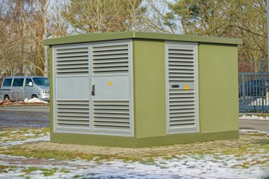 Çevresinde kar ve çimen bulunan asfalt bir alanda metal havalandırma panelleri ve uyarı levhaları olan yeşil elektrik trafo istasyonu..