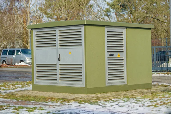 Çevresinde kar ve çimen bulunan asfalt bir alanda metal havalandırma panelleri ve uyarı levhaları olan yeşil elektrik trafo istasyonu..