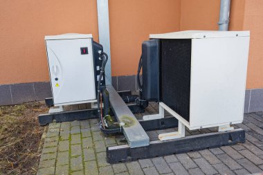 Beyaz klima ve elektrik kontrol kutusu, turuncu duvar arka planı ve bağlı boruları olan asfalt bir yüzeye monte edilmiş..