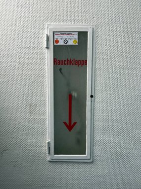 Duman kontrol paneli, binalarda duman havalandırması için gerekli güvenlik ekipmanlarını gösteren, dokulu beyaz bir duvarda kırmızı bir ok ile Almanca etiketlenmiş..