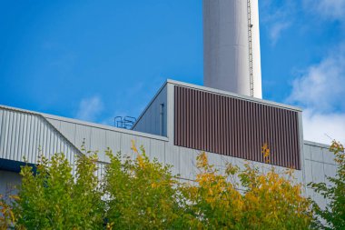 Stadtwerke Flensburg adında büyük bir sanayi binası, üzerinde beyaz bir egzoz yığını yükselen açık mavi bir gökyüzünün altında duruyor. Penceresiz cephe üretim, depolama veya işleme öneriyor.