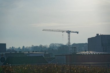 Tipik bir şehir manzarası katmanlı çatıları ve apartman binalarını bir arada gösteriyor. Ufuk çizgisinden yükselen uzak bir vinç bölgede devam eden inşaat çalışmalarını gösteriyor..