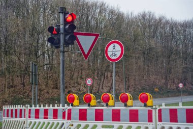 Kırmızı bir trafik ışığı araçları durdururken, bir karayolu bölümü geçici inşaat çitleri tarafından engelleniyor. Kurulum kontrollü yol çalışmalarını ve alternatif trafik geçitlerini gösteriyor.