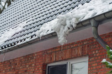 Bir kırmızı tuğla cephesinde havalandırma için bir pencere açılır ve çatıdan bir miktar kar sarkır. Tek bir buz sarkıtı aşağıya doğru düşer ve dondurucu koşulları vurgular..
