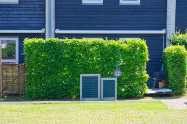 Kırpılmış yeşil bir çit, örülmüş ahşap bir kapının yanında duran iki metal paneli olan koyu renkli bir bina duvarını çiziyor. Sahne sessiz ve kullanışlı geliyor, düzgün kenarları ve küçük detayları var..