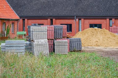 Gri ve kırmızıyla kaplı bir kum yığını ve kaldırım blokları yeni bir evin yanında duruyor. Depolanan malzemeler arazinin çevresindeki peyzaj ya da sokak onarımına işaret ediyor..