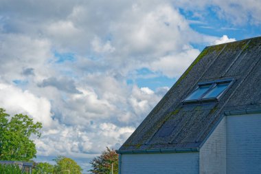 Beyaz boyalı bir tuğla ev, yeşil yosunlarla kaplı eski bir çatı ve havalandırma için açık bir çift pencere gösteriyor. Çatının arkasında yumuşak beyaz bulutlar sürükleniyor..