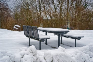 Metal piknik masası ve karla kaplı banklar sessiz bir kış parkında çıplak ağaçlar ve yerde kar yığınlarıyla çevrili..