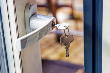 Üzerinde anahtarlar olan gümüş bir kapı kolu kısmen açık bir kapının kilidine yerleştirilmiş, sıcak tonlu bulanık bir arka plan gösteriliyor..