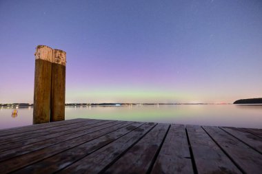 Huzurlu bir ahşap iskele, sakin bir göle uzanıyor. Yıldızlı gece gökyüzünü ve ufuktaki uzak ışıklarla soluk yeşil bir aurora yansıtıyor..