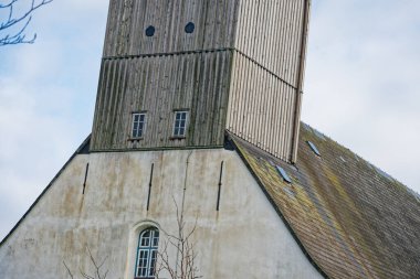 Dikey ahşap kaplamalı yaşlı kilise kulesi, küçük pencereler ve kırsal alanda bulutlu bir gökyüzü arka planına karşı yosun kaplı kiremitli çatı.