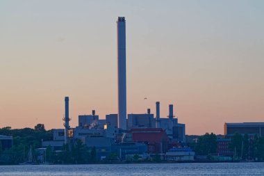 Gün batımında açık gökyüzü ve etrafını çevreleyen ağaçlarla birlikte bir su kütlesinin yanında birden fazla bacası olan büyük bir sanayi enerji santralinin görüntüsü..