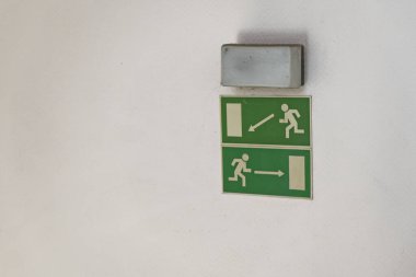 Beyaz bir duvarda acil durum ya da yangın durumunda güvenli tahliye rotalarını gösteren iki yeşil çıkış işareti var..