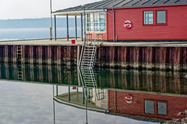 Rıhtımın sakin suyuna yansıyan can yeleği ve metal merdiveni olan kırmızı ahşap bir kayıkhane içeren huzurlu bir liman..