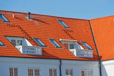 Parlak turuncu kiremitli çatı, çoklu çatı pencereleri ve açık mavi gökyüzünün altında iki küçük beyaz balkon..
