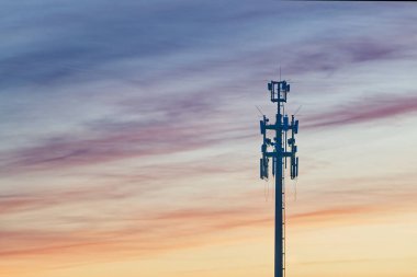 Telekomünikasyon kulesi, yumuşak pastel bulutlar ve renklerin yer aldığı canlı bir günbatımı gökyüzüne karşı çoklu antenlerle dimdik ayakta duruyor..