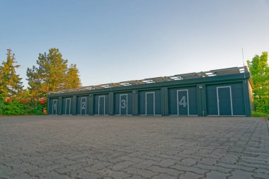 Çağdaş depolama birimlerinin geniş sayılı kapıları, temiz mavi gökyüzünün altındaki ağaçlarla çevrili asfalt zemine kurulmuş..