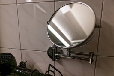 Krom ayarlanabilir kollu yuvarlak bir banyo aynası fayansla döşenmiş siyah bir saç kurutma makinesinin yanında ışığı yansıtan ve banyonun iç kısımlarını yansıtan..