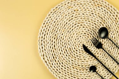 Kahverengi hasır masa paspası, sarı arka planda siyah çatal bıçak takımı. Saman yuvarlak tepsi, düz konum, kopyalama alanı. 