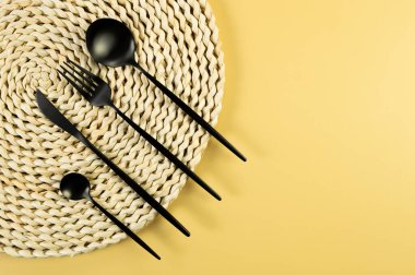 Kahverengi hasır masa paspası, sarı arka planda siyah çatal bıçak takımı. Saman yuvarlak tepsi, düz konum, kopyalama alanı. 