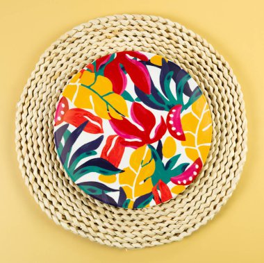 Kahverengi hasır masa paspasının üst görüntüsü, sarı arka planda renkli plaka. Saman tepsisi, egzotik bitki tabağı, yaz pikniği. Düz yat, boşluğu kopyala. 