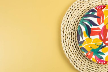 Kahverengi hasır masa paspasının üst görüntüsü, sarı arka planda renkli plaka. Saman tepsisi, egzotik bitki tabağı, yaz pikniği. Düz yat, boşluğu kopyala. 