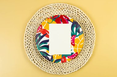 Kahverengi hasır masa paspası, beyaz kart, sarı arka planda renkli plaka. Saman tepsisi, egzotik bitki tabağı, yaz pikniği. Düz yat, boşluğu kopyala. 