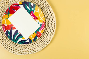 Kahverengi hasır masa paspası, beyaz kart, sarı arka planda renkli plaka. Saman tepsisi, egzotik bitki tabağı, yaz pikniği. Düz yat, boşluğu kopyala. 