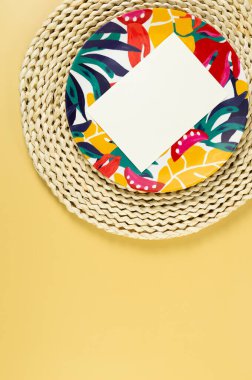 Kahverengi hasır masa paspası, beyaz kart, sarı arka planda renkli plaka. Saman tepsisi, egzotik bitki tabağı, yaz pikniği. Düz yat, boşluğu kopyala. 