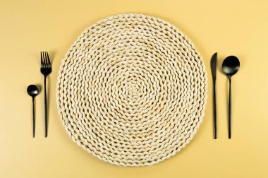 Kahverengi hasır masa paspası, sarı arka planda siyah çatal bıçak takımı. Saman yuvarlak tepsi, düz konum, kopyalama alanı. 