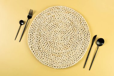 Kahverengi hasır masa paspası, sarı arka planda siyah çatal bıçak takımı. Saman yuvarlak tepsi, düz konum, kopyalama alanı. 