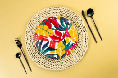 Kahverengi hasır masa paspası, renkli tabak, sarı arka planda siyah çatal bıçak takımı. Saman tepsisi, egzotik bitki tabağı, yaz pikniği. Düz yat, boşluğu kopyala. 