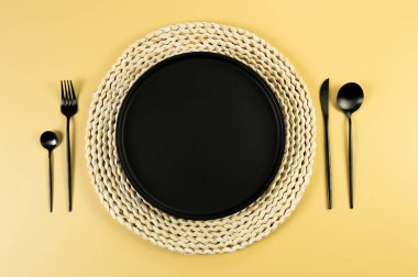 Kahverengi hasır masa paspası, siyah tabak, sarı arka planda siyah çatal bıçak takımı. Saman yuvarlak tepsi, düz konum, kopyalama alanı. 