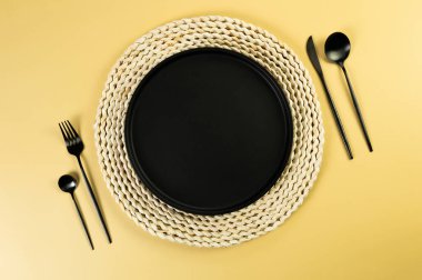 Kahverengi hasır masa paspası, siyah tabak, sarı arka planda siyah çatal bıçak takımı. Saman yuvarlak tepsi, düz konum, kopyalama alanı. 