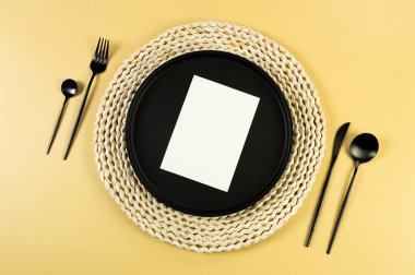 Kahverengi hasır masa paspası, siyah tabak, sarı arka planda siyah çatal bıçak takımı. Beyaz kart, saman yuvarlak tepsi, düz konum, kopyalama alanı. 