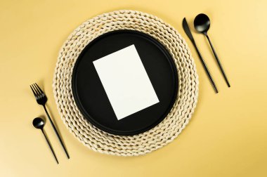 Kahverengi hasır masa paspası, siyah tabak, sarı arka planda siyah çatal bıçak takımı. Beyaz kart, saman yuvarlak tepsi, düz konum, kopyalama alanı. 