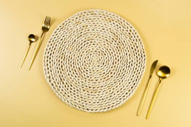 Kahverengi hasır masa paspası, sarı arka planda altın çatal bıçak seti. Saman yuvarlak tepsi, düz konum, kopyalama alanı. 