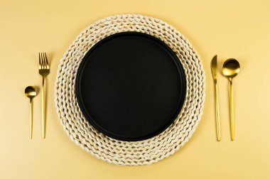 Kahverengi hasır masa paspası, siyah tabak, sarı arka planda altın çatal bıçak takımı. Saman yuvarlak tepsi, düz konum, kopyalama alanı. 