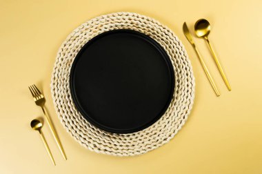 Kahverengi hasır masa paspası, siyah tabak, sarı arka planda altın çatal bıçak takımı. Saman yuvarlak tepsi, düz konum, kopyalama alanı. 