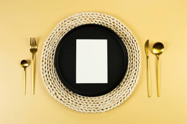 Kahverengi hasır masa paspası, siyah tabak, sarı arka planda altın çatal bıçak takımı. Beyaz kart, saman yuvarlak tepsi, düz konum, kopyalama alanı. 