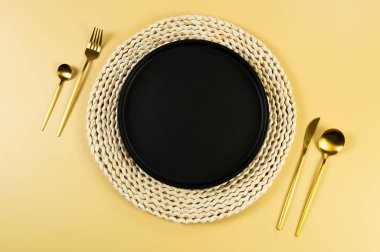 Kahverengi hasır masa paspası, siyah tabak, sarı arka planda altın çatal bıçak takımı. Saman yuvarlak tepsi, düz konum, kopyalama alanı. 