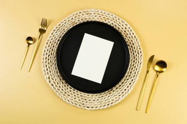 Kahverengi hasır masa paspası, siyah tabak, sarı arka planda altın çatal bıçak takımı. Beyaz kart, saman yuvarlak tepsi, düz konum, kopyalama alanı. 