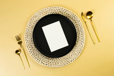 Kahverengi hasır masa paspası, siyah tabak, sarı arka planda altın çatal bıçak takımı. Beyaz kart, saman yuvarlak tepsi, düz konum, kopyalama alanı. 