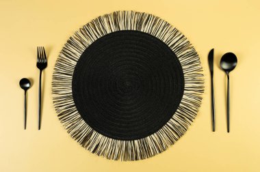 Siyah hasır masa paspası, sarı arka planda siyah çatal bıçak takımı. Saman yuvarlak tepsi, düz konum, kopyalama alanı. 