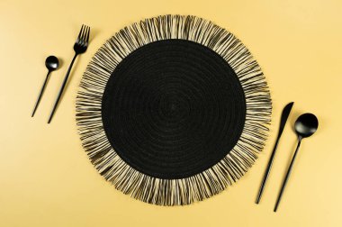 Siyah hasır masa paspası, sarı arka planda siyah çatal bıçak takımı. Saman yuvarlak tepsi, düz konum, kopyalama alanı. 