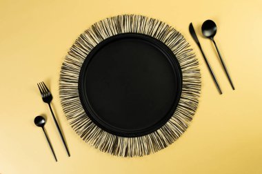 Siyah hasır masa paspası, sarı arka planda siyah çatal bıçak takımı. Saman yuvarlak tepsi, düz konum, kopyalama alanı. 