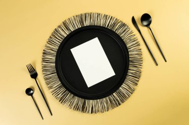 Siyah hasır masa paspası, siyah çatal bıçak takımı, sarı arka planda beyaz kart. Saman yuvarlak tepsi, düz konum, kopyalama alanı. 