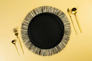 Siyah hasır masa paspası, siyah tabak, sarı arka planda altın çatal bıçak. Saman yuvarlak tepsi, düz konum, kopyalama alanı. 