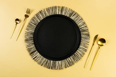 Siyah hasır masa paspası, siyah tabak, sarı arka planda altın çatal bıçak. Saman yuvarlak tepsi, düz konum, kopyalama alanı. 