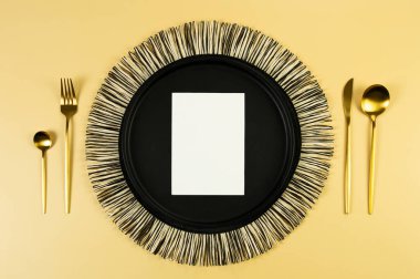 Siyah hasır masa paspası, siyah tabak, altın çatal bıçak takımı, sarı arka planda beyaz kart. Saman yuvarlak tepsi, düz konum, kopyalama alanı. 